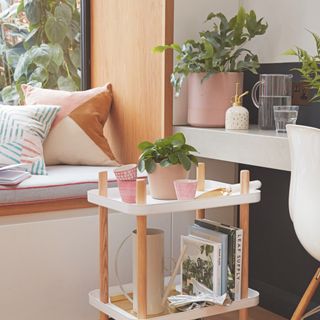 corner of living room, window seat, pastel cushions, wood and white storage unit with plant and cups, plant on side with jug or water and glass