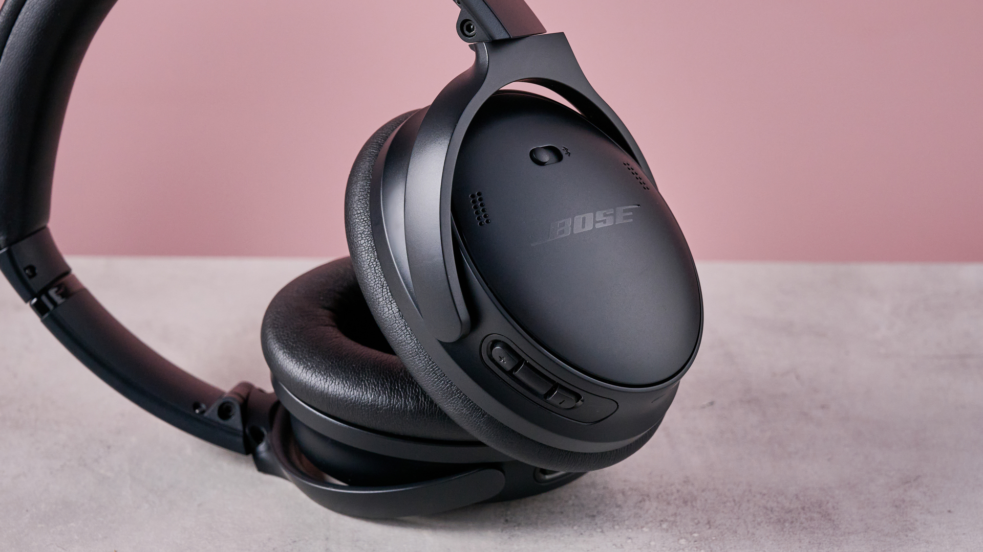 Bose QuietComfort Headphones on a gray surface with a pink background, showing the controls on the earcup