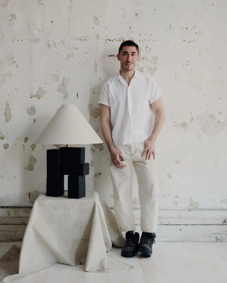 A young man dressed in a white short-sleeved shirt, beige trousers, and black shoes stands against a rough creme wall next to a table lamp in the same color tones.