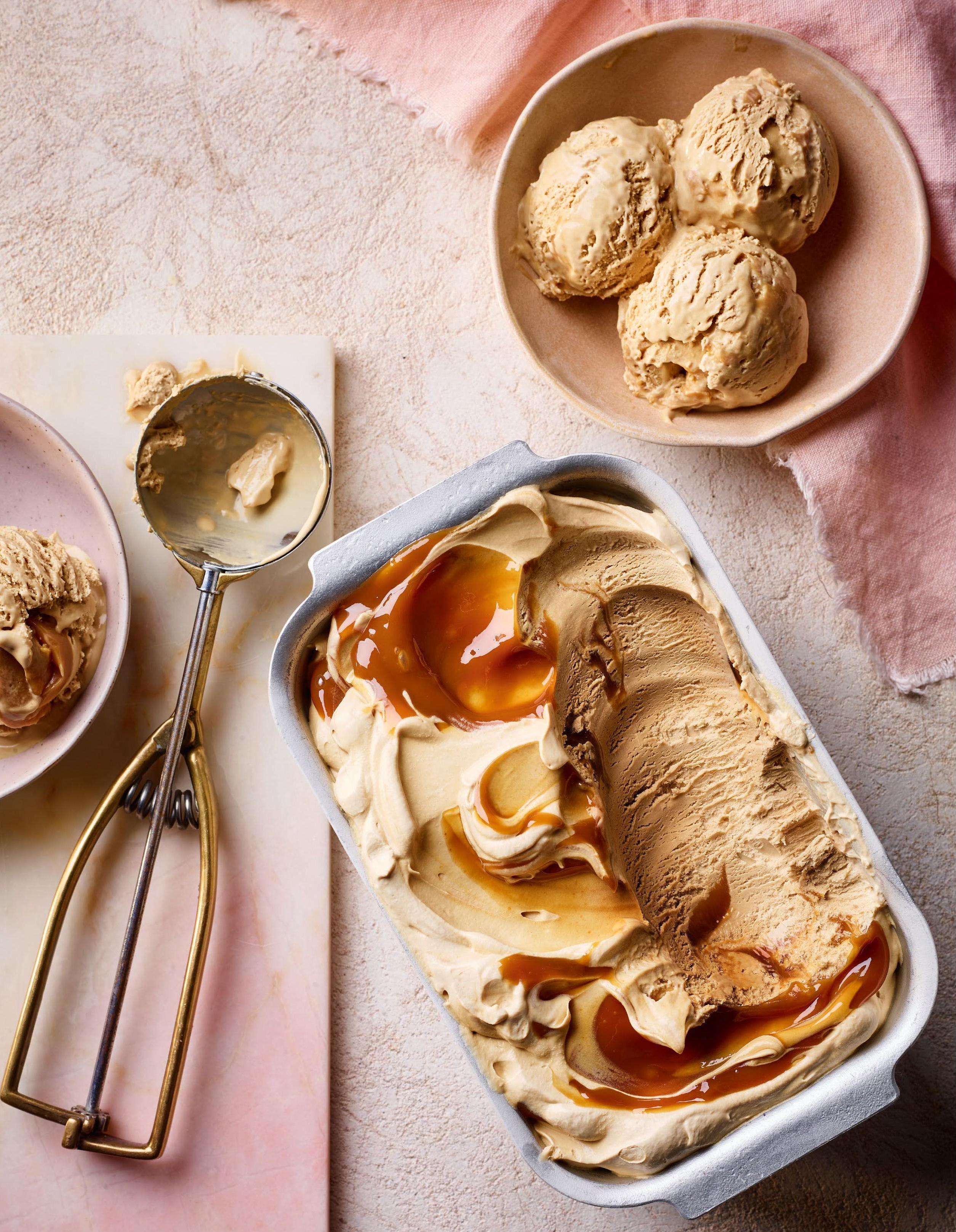 A creamy coffee ice cream in a rectangular dish, swirled with caramel, accompanied by three scoops in a small bowl and an ice cream scoop on a light textured surface.