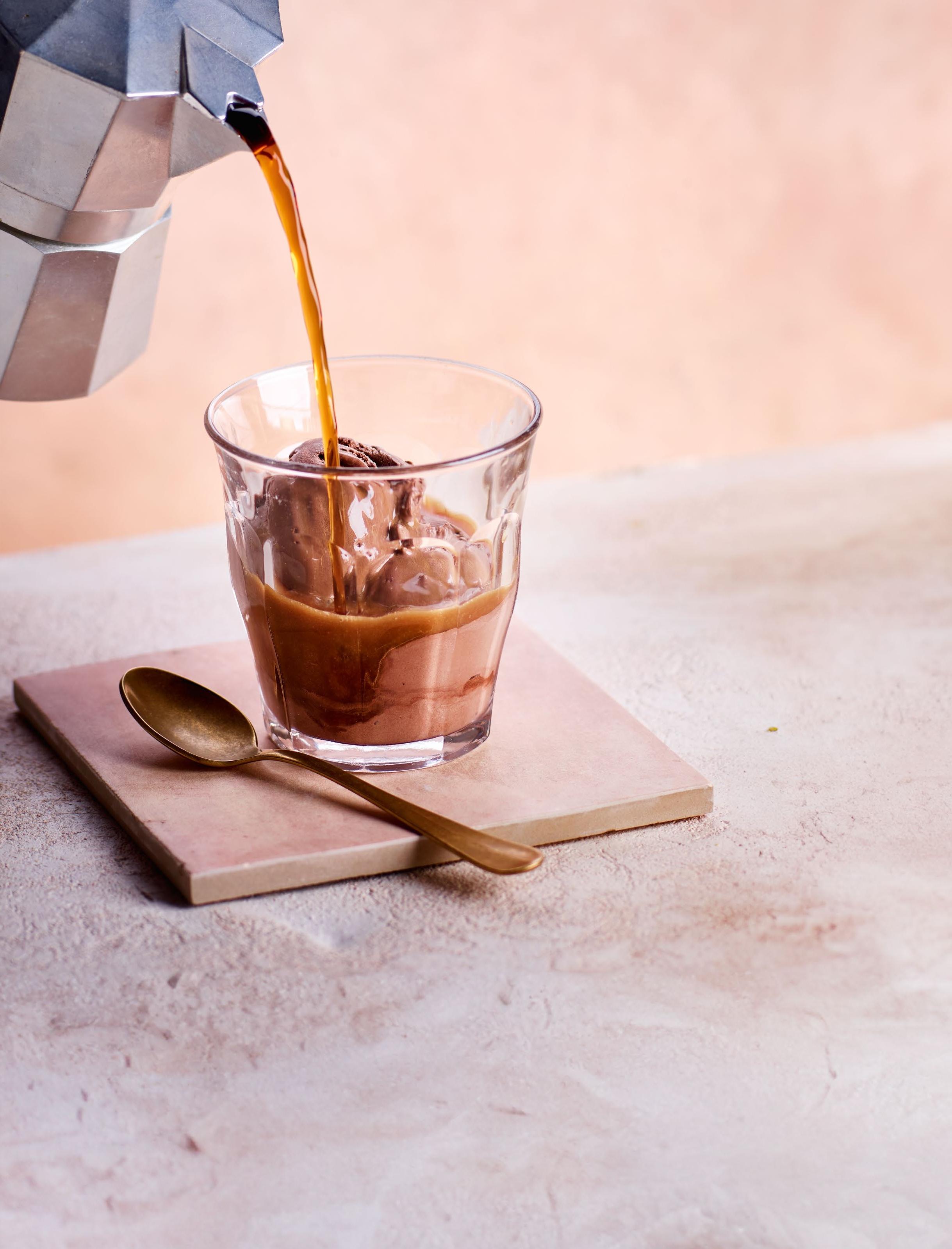 A glass of ice cream being topped with freshly brewed coffee, set on a light-colored surface with a spoon beside it.