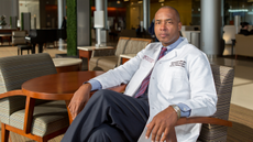 A man in a white doctor's coat sits in a chair and looks at the camera