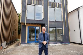 Kevin McCloud stood outside of the house featured on the new episode of Grand Designs: The Streets shows an incomplete wooden framed, large-windowed self build 