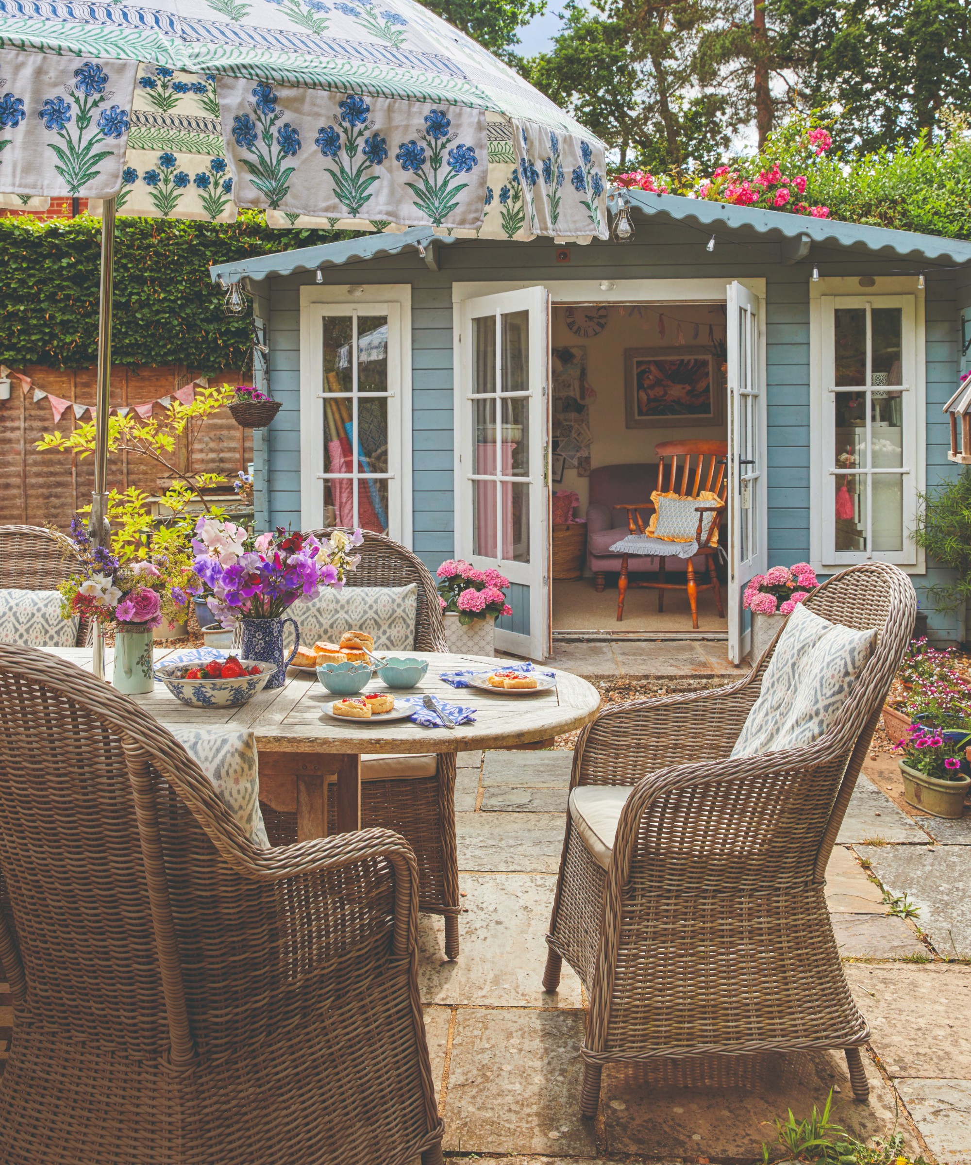 A patio with a wooden dining tale and rattan dining chairs and a floral-print parasol