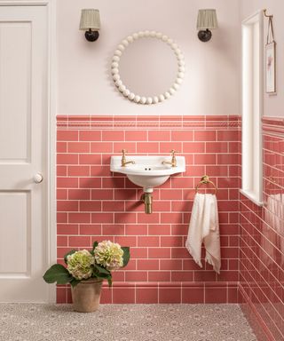 Dusky pink subway wall tiles in a small bathroom with classic looking wall sconces, a round bobbled edge white mirror, white ink with brushed gold hardware, and flowers