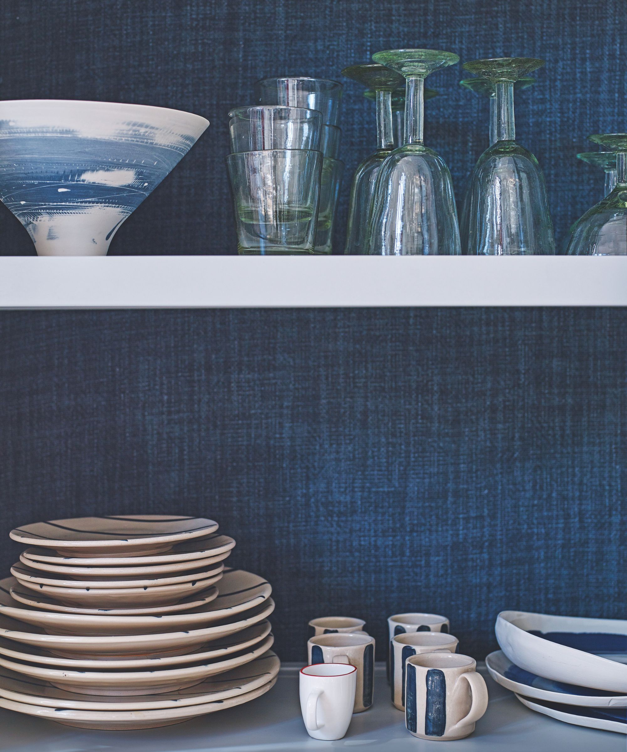 Stacks of white and blue plates and mugs on white open shelving with a blue background. On the shelf above are green glasses and a white and blue bowl.