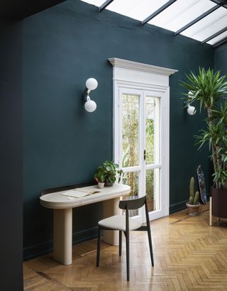 moody open plan space with simple desk set up, wooden floor, and white doors out to the garden to the right of the desk