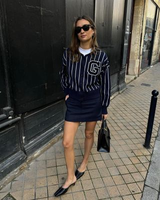 Woman wearing striped jersey, navy miniskirt, black heeled mules, and black sunglasses standing on brick street.