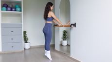 A woman stands at a bar in a studio doing calf raises. She is on her tip toes. In front of her is a mirror, reflecting the green leafy plant behind her. We also see a storage unit, stocked with Pilates balls.