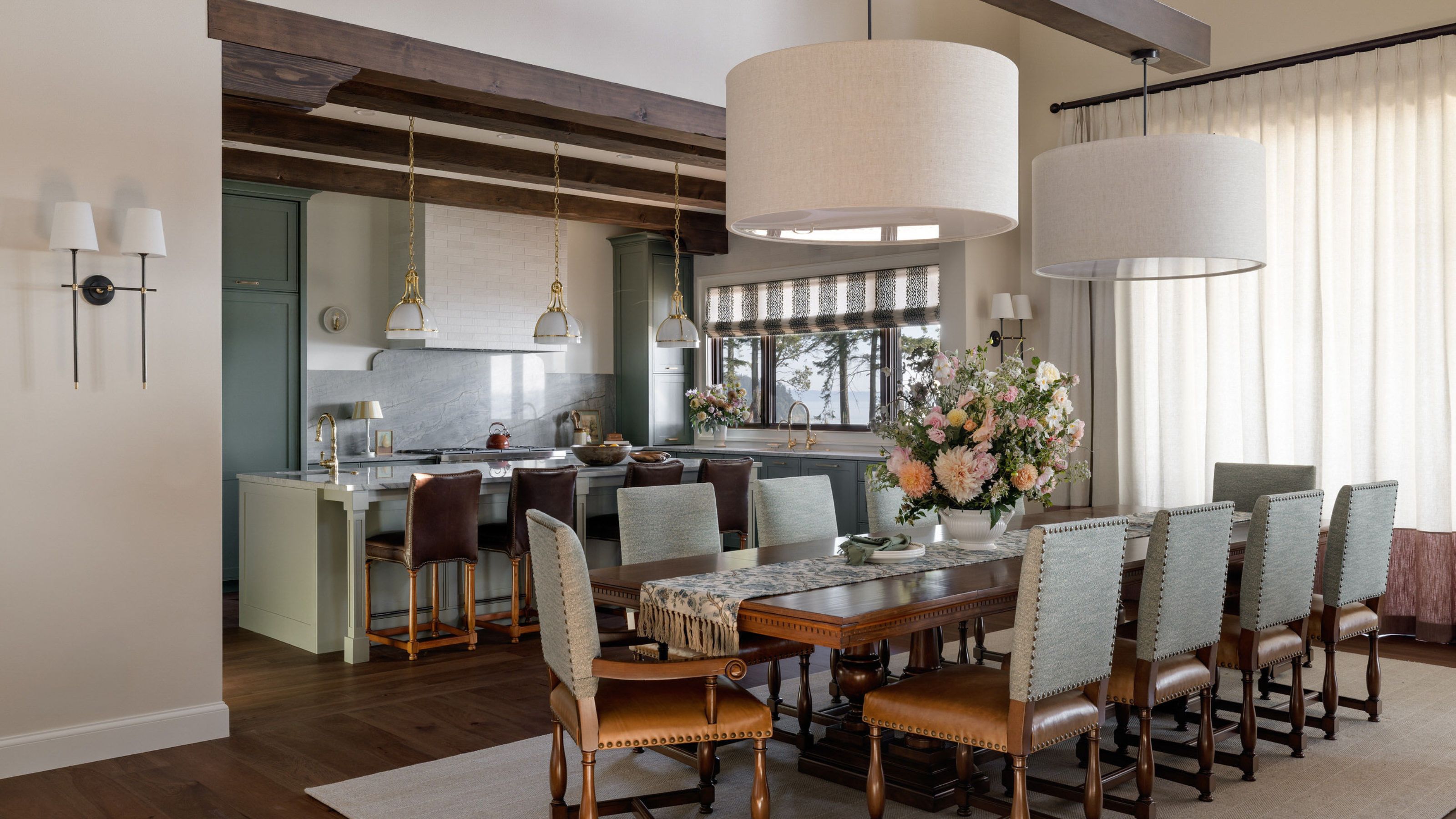 An open-plan kitchen and dining room with wooden flooring, a large dining table and leather chairs, muted green kitchen cabinets, and wood ceiling beams.