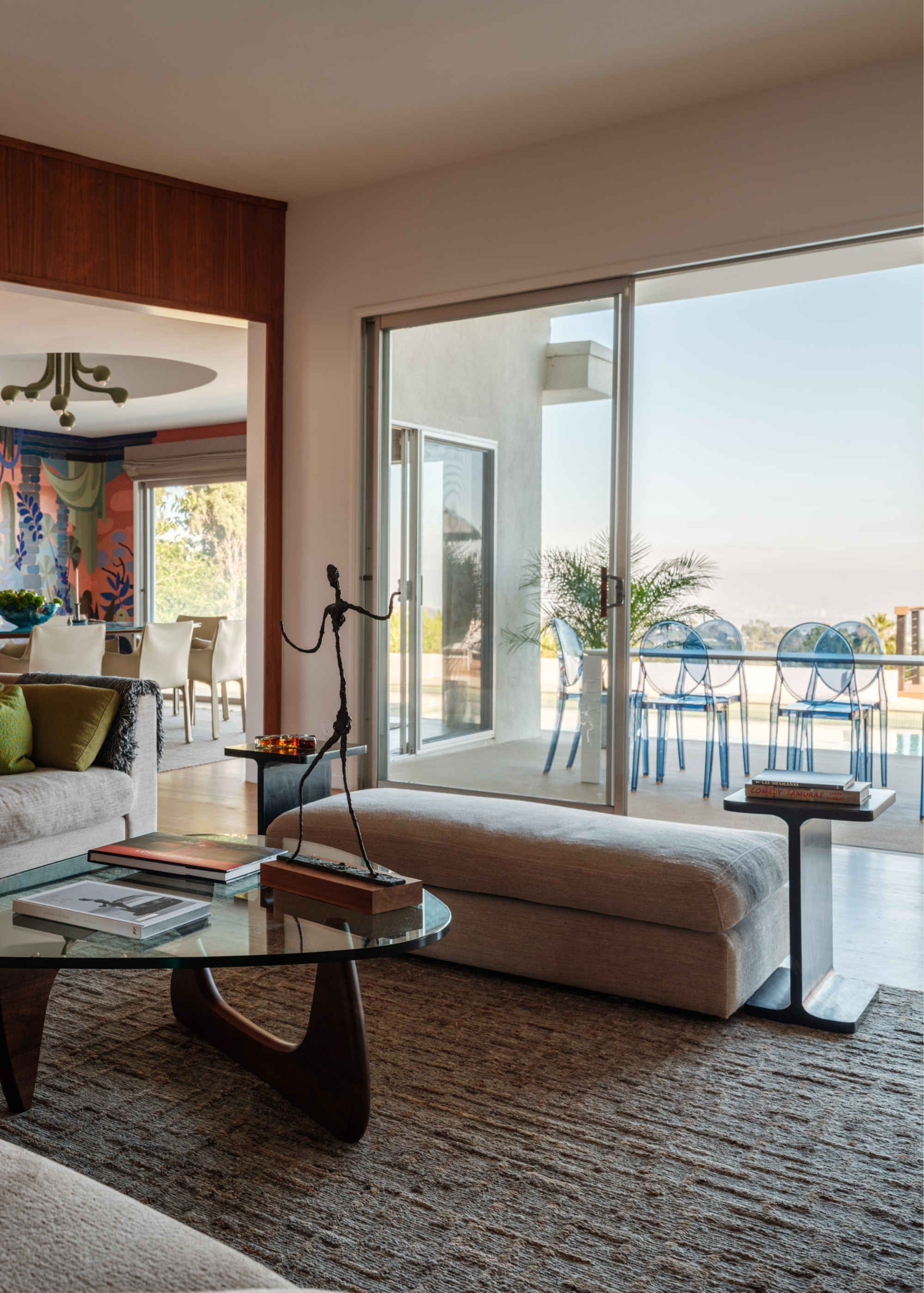 A living room with a glass door leading to an outdoor dining space with translucent blue chairs. In the living room is a textured area rug, a glass coffee table with books, a statue, and a beige sofa bench