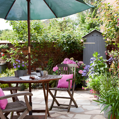 Small, slim garden shed bind a wood dining table with pink cushions and green parasol.