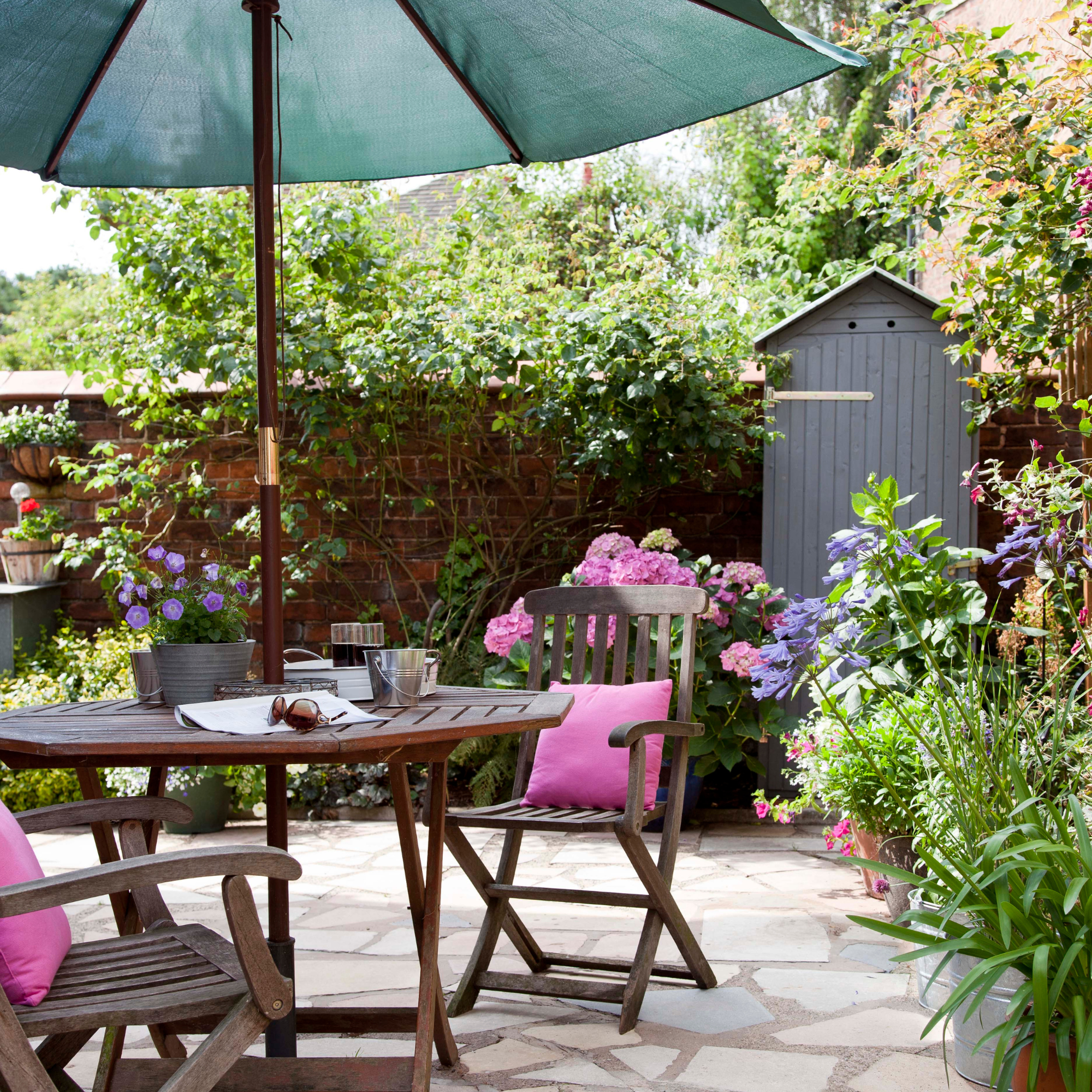 Small, slim garden shed bind a wood dining table with pink cushions and green parasol.