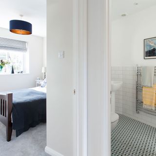 patterned floor bathroom with a wall-mounted towel rack and a sink, next to a bedroom weith a wooden bed and a navy bed-throw and a navy ceiling lamp with a window