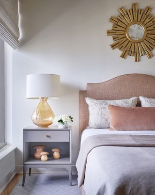 white bedroom with gray side table and rug, coral headboard and cushions, sunburst mirror, amber glass based table lamp