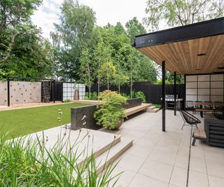 A newly landscaped back garden with light coloured paving and covered patio with children's play space in the back