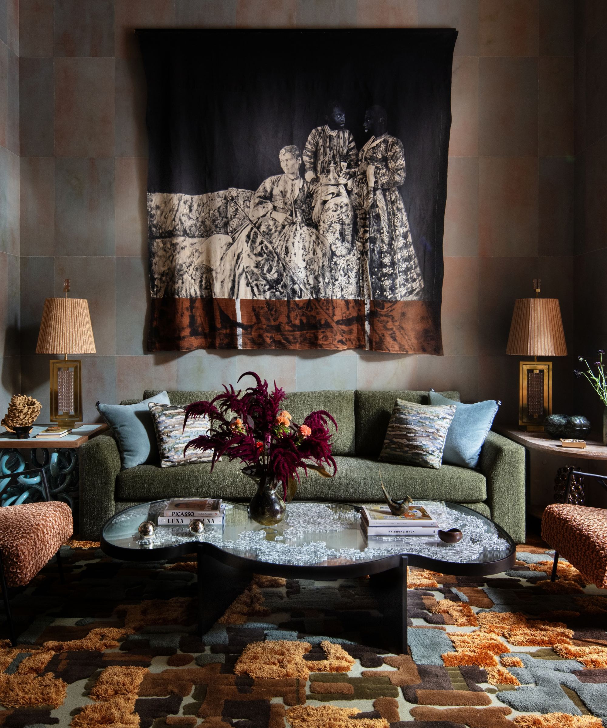 Moody, eclectic NYC living room featuring a jewel-toned abstract high-low rug, sculptural coffee table, moss-green couch with square throw pillows, and a large tapestry hanging overhead.