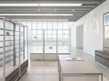 Interior view at the Turner Contemporary shop featuring white walls, windows, light grey flooring, white and grey shelving units with various items on display, grey tables and drawers, a neon sign of a book, framed wall art and rectangular ceiling lights