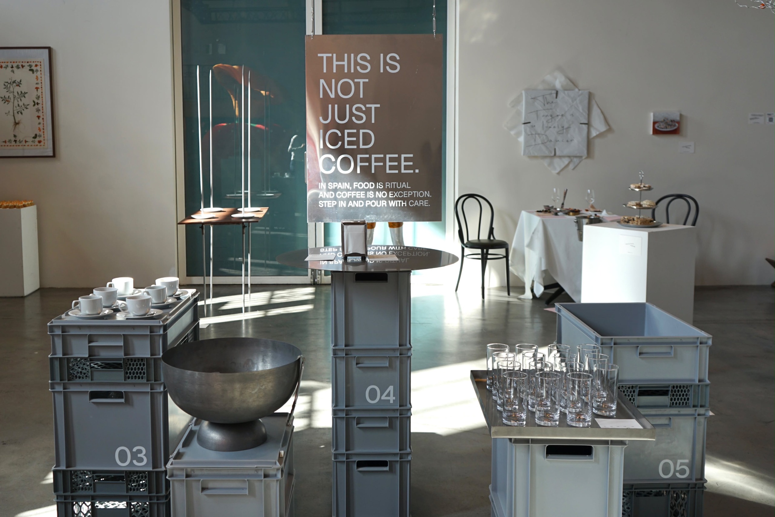 Installation view of an exhibition featuring industrial boxes, cups, flasses, and furniture, and a banner saying "this is not just iced coffee."