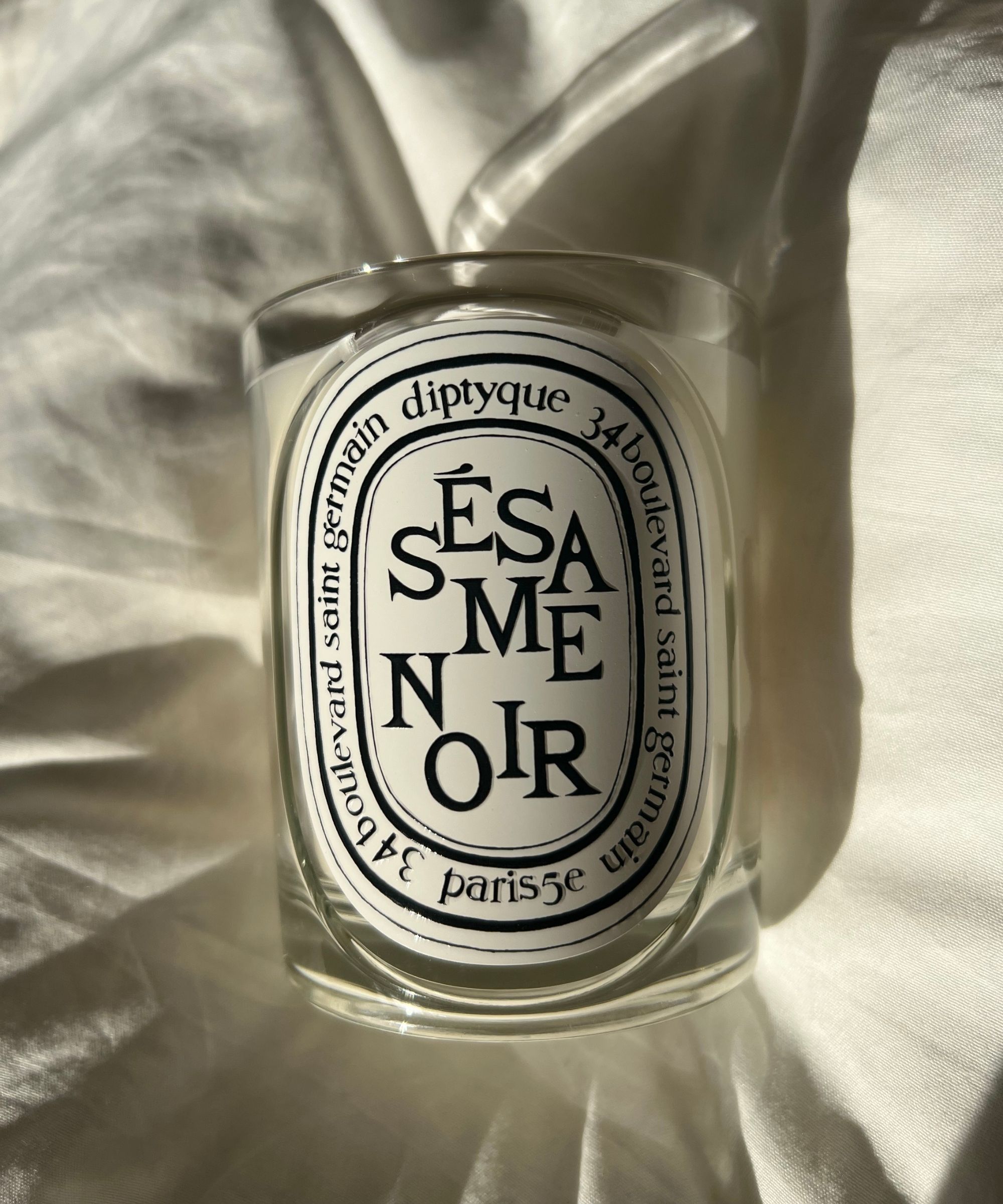 Diptyque Sesame Noir candle in a glass vessel with a white oval label and black lettering pictured in natural sunlight on white linen bedding.