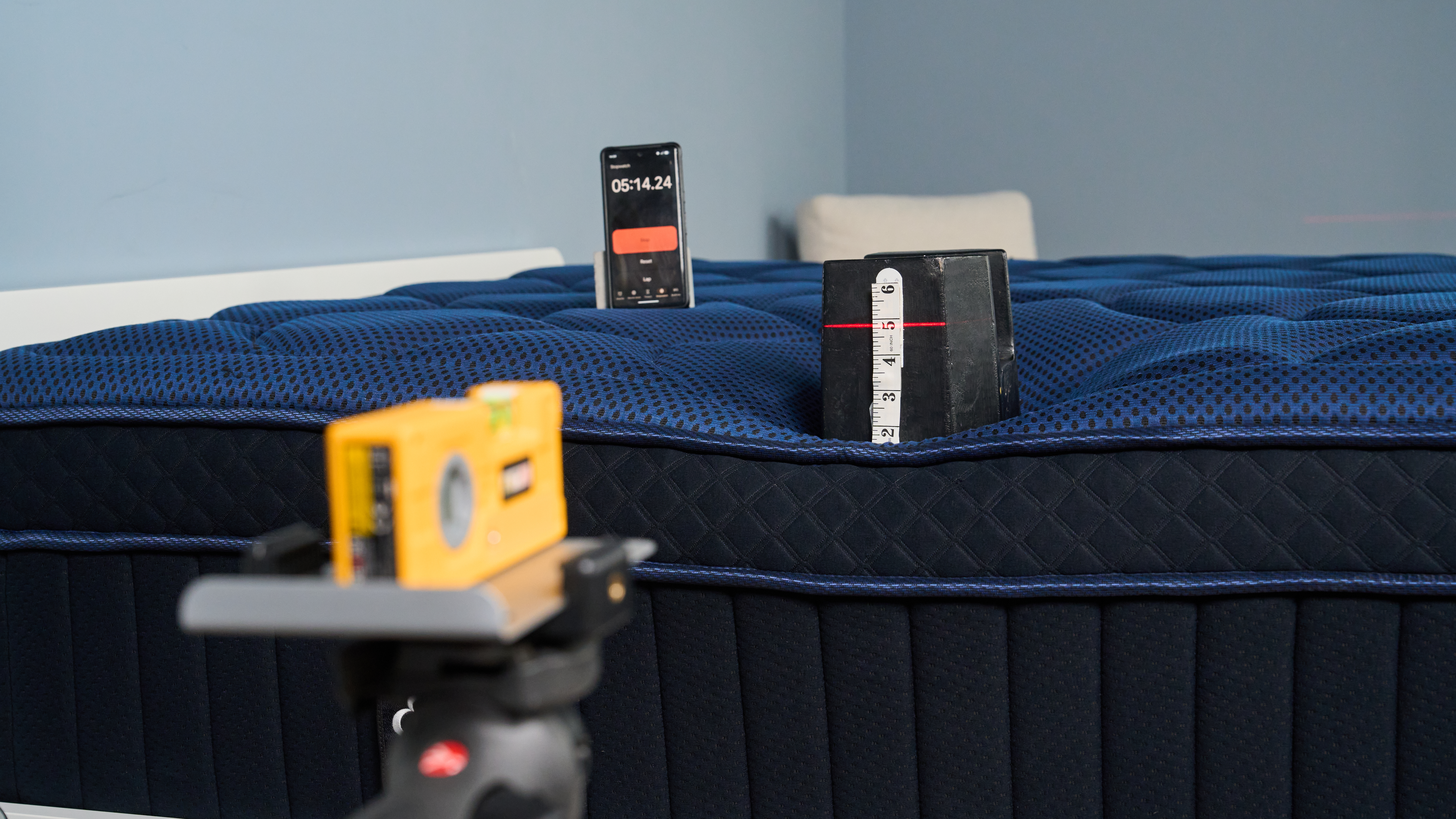 The image shows a mattress tester wearing a pink shirt and blue sweater vest place a 25kg weight on the base and side of a mattress to determine its edge support