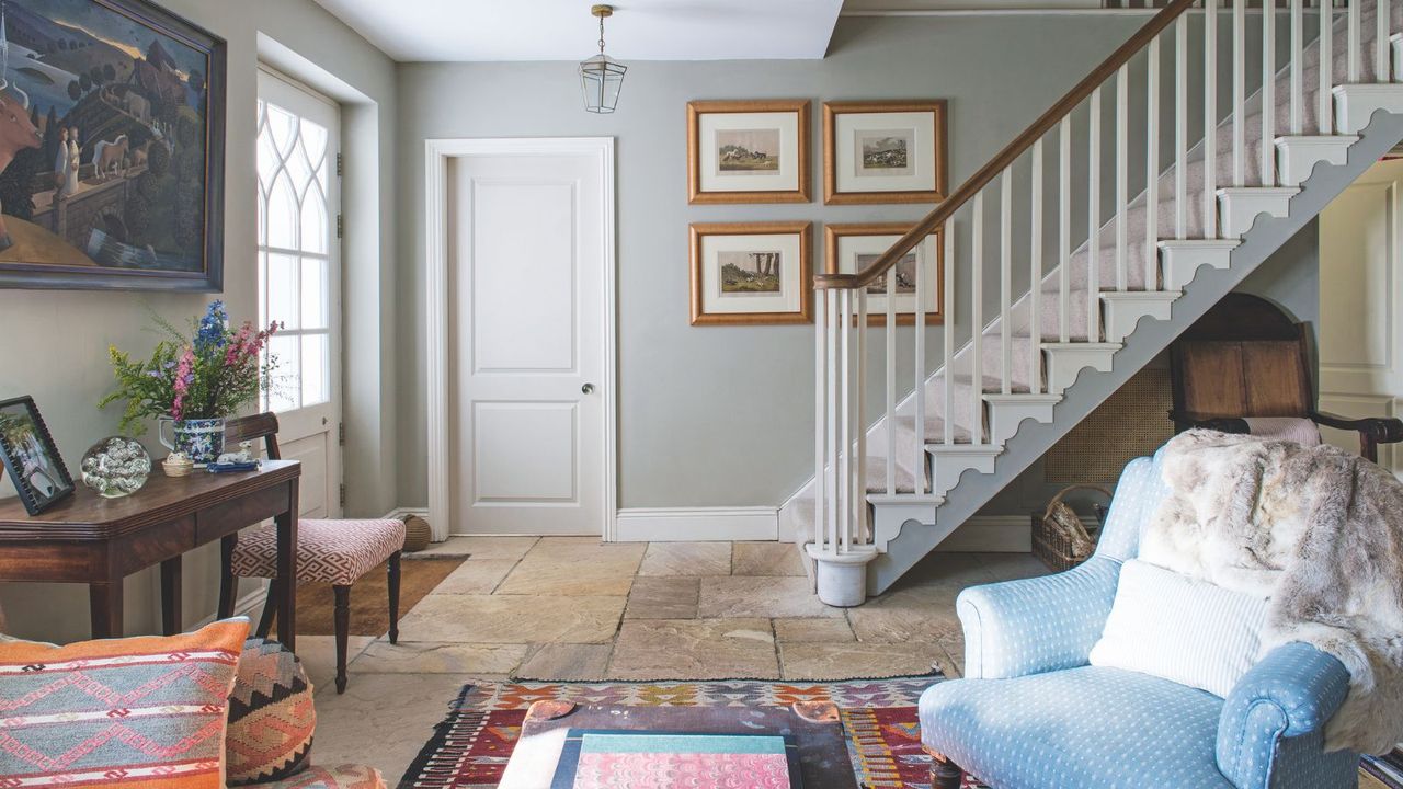 A bright and colorful beige tiled entryway with grey green walls, a statement patterned rug and matching pillows, a blue armchair with a fur blanket, a wooden console table with flowers and photographs, and gold framed wall art and paintings with white stairs in the background next to a white door