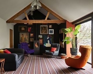 Wood burner in living room with various velvet chairs in purple and orange, striped carpeted floor and framed wall prints