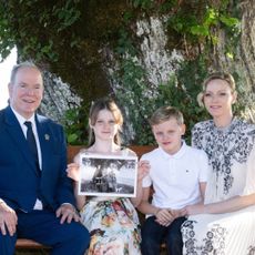 Prince Albert, Princess Charlene and their twins sitting in front of a tree holding a black and white photo