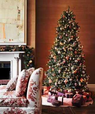 A tall decorated real pine Christmas tree by a traditional fireplace, and a garland laid on the mantlepiece, a flora patterned sofa and rich brown painted walls.