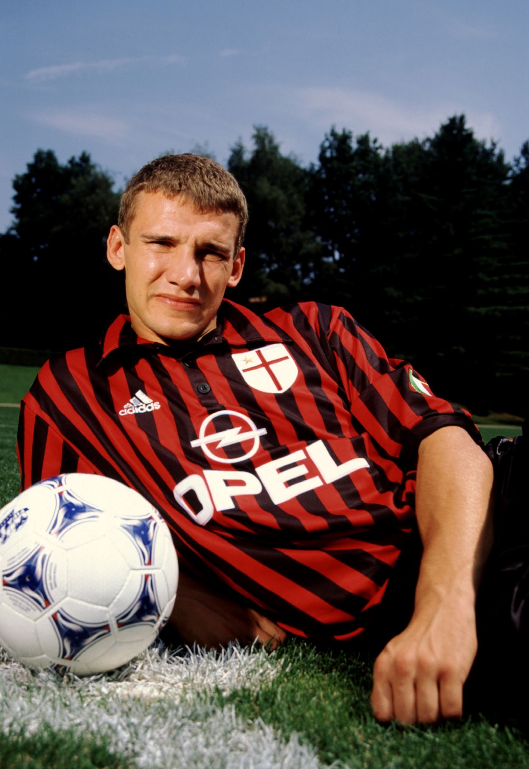 A football player in a red and black striped jersey lying on the grass next to a soccer ball, reflecting on his childhood dream of winning the Ballon d'Or.