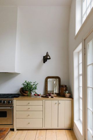 minimalist kitchen with soft butter yellow painted cabinets, with a mirror, vase, fruit, and potted plant styled on counter, and a wall sconce hanging above