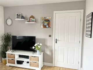 A grey feature wall decorated with art, shelves, photos, a clock, and in front of a white TV stand with a black TV and some white tulips.