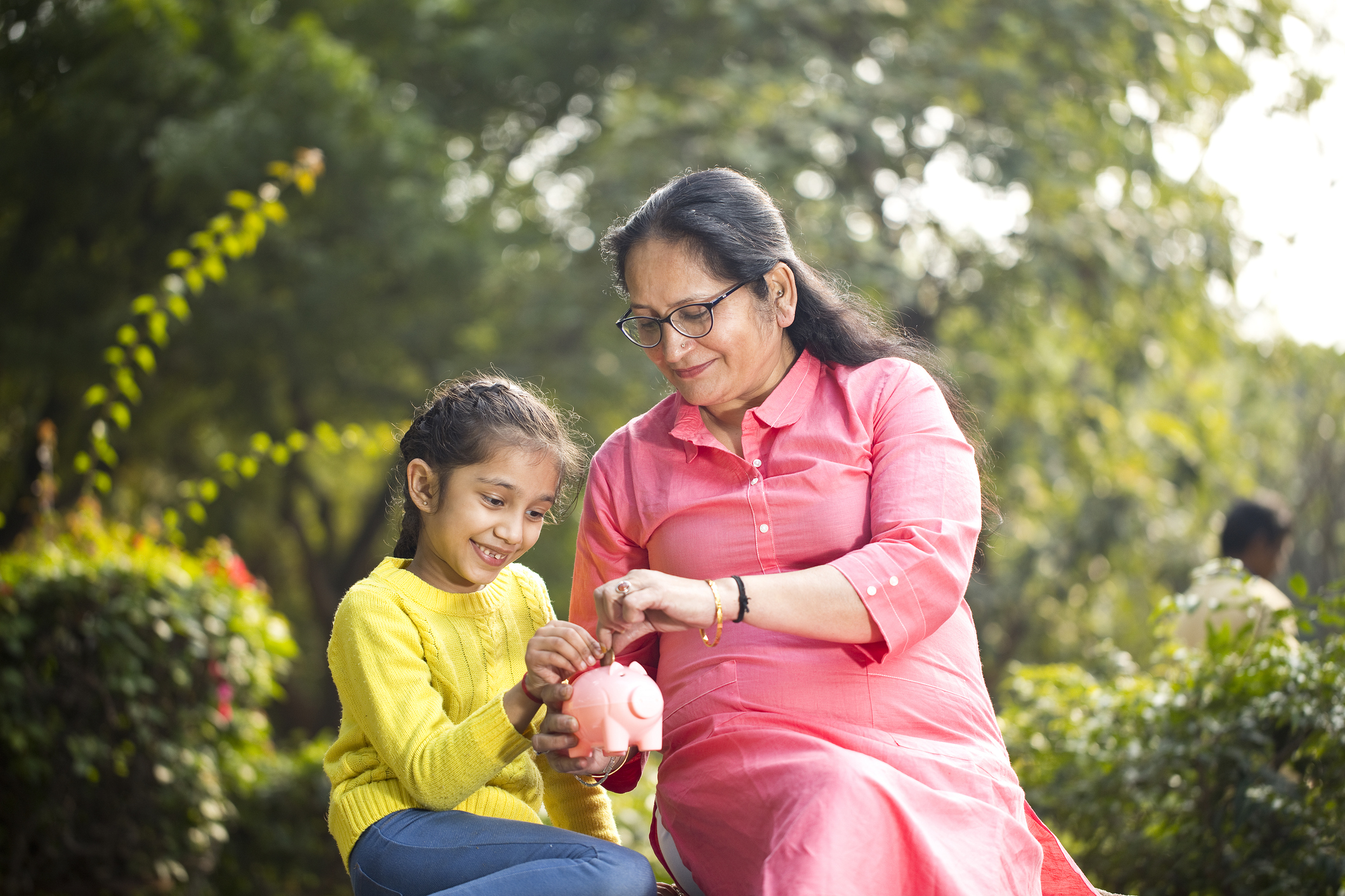Grandmother with granddaughter in a park, putting money in piggy bank