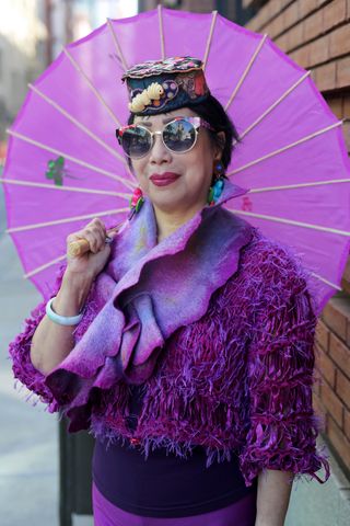 A photo of Terri Wong in an over 60 outfit featuring a purple top, purple knit sweater, purple scarf, sunglasses, hat, and holding a purple parasol.