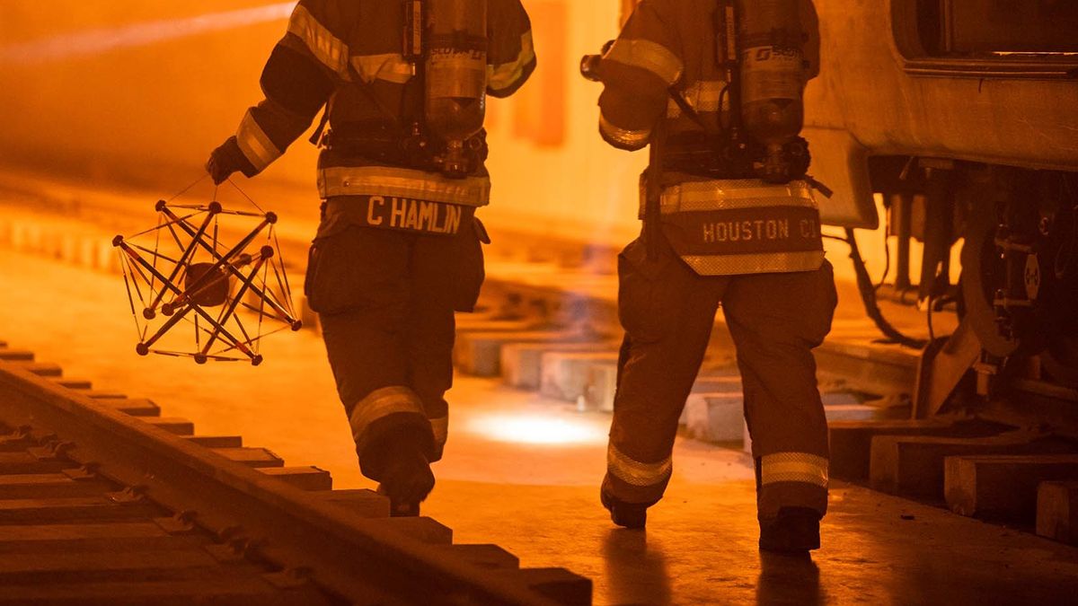 Two 1st responders are seen in an orange-hued scene. The one on the left is holding the squishy robot which looks like a sphere trapped inside a multi-pronged frame.