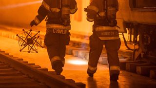 Two 1st responders are seen in an orange-hued scene. The one on the left is holding the squishy robot which looks like a sphere trapped inside a multi-pronged frame.