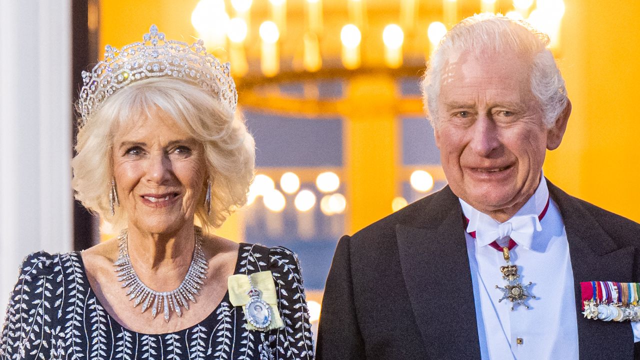 Queen Camilla wears a sparkling tiara and a black dress with a white floral design, while King Charles wears a formal suit with medals and a red sash