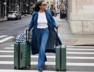 Woman in denim trench and jeans rolling green suitcases on a New York City street