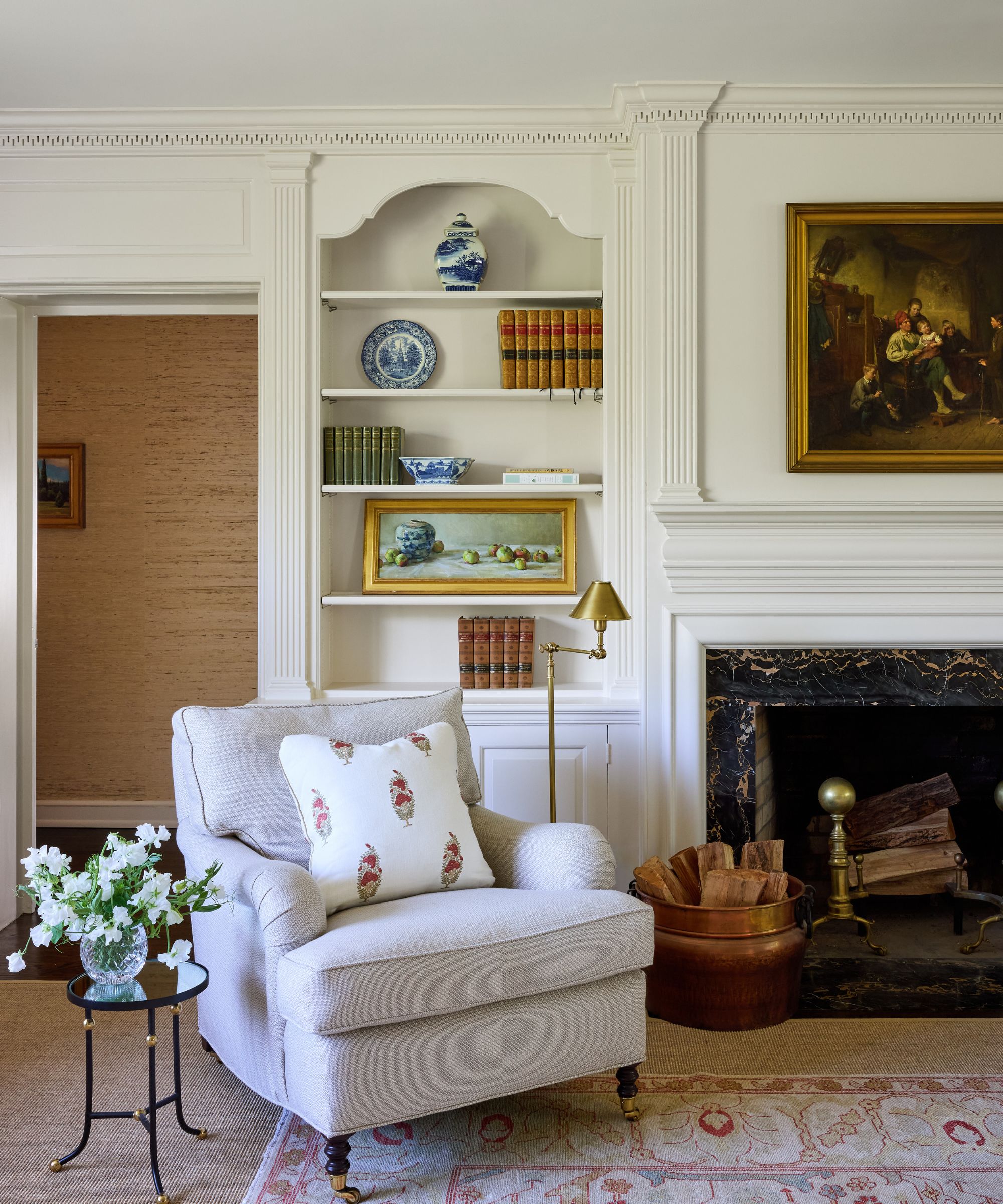 Traditional living room with crown molding, painting above fireplace, marble fireplace, cream armchair and paisley pillow and bookshelf with books and china