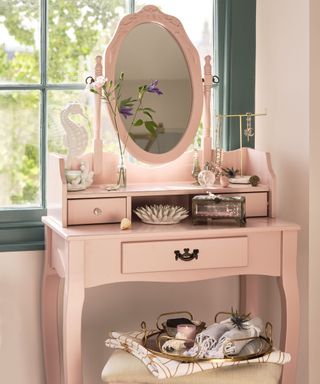 A light pink vanity with a mirror, flowers, a gold jewellery holder, and a jewellery box on it, with a white stool underneath with a gold decorated tray and a dark green window behind it
