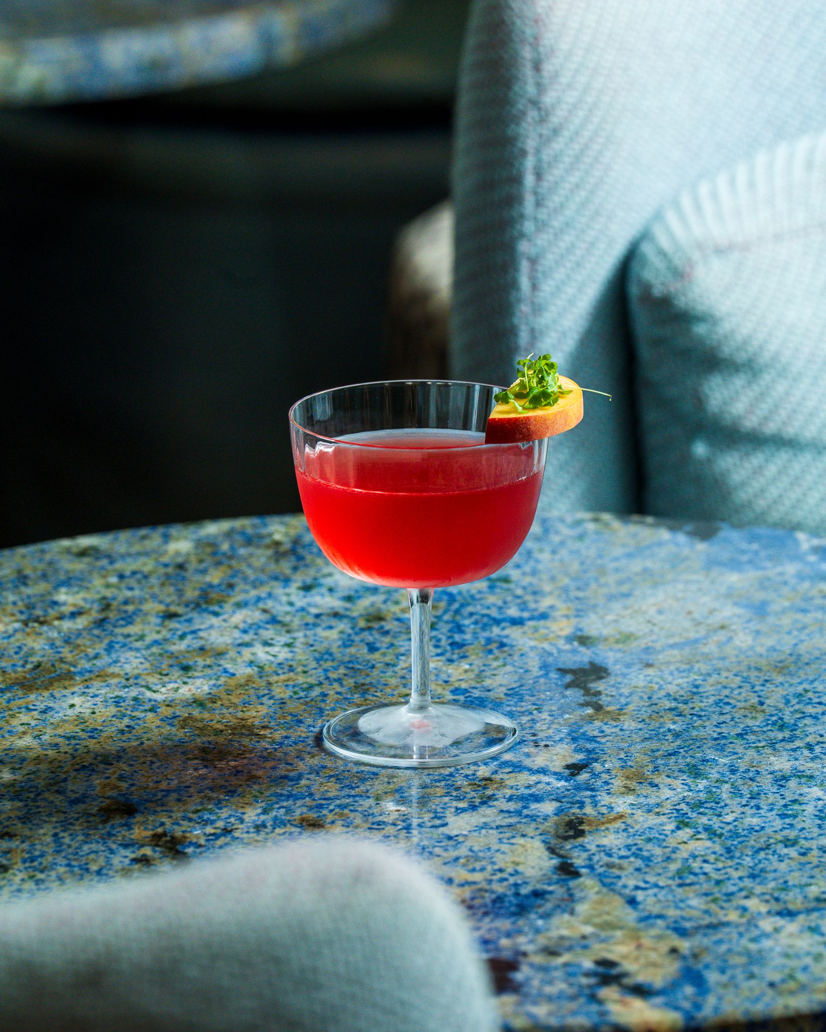 Two red cocktails served in transparent glasses sitting on terrazzo green, or blue, tables, respectively.