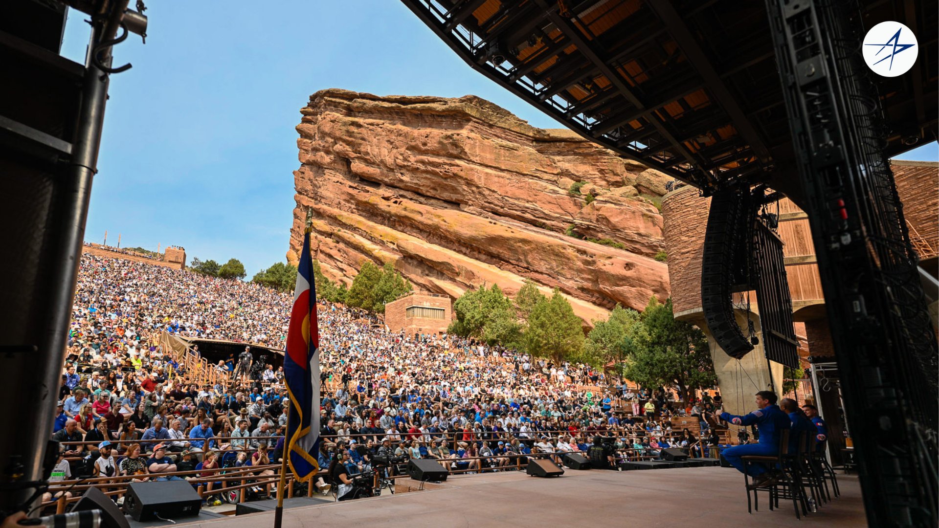 four people in blue flight suits stand on a stage in an amphitheater surrounded by red stone cliffs