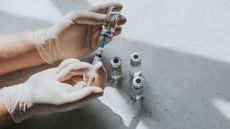 gloves hands drawing liquid from a vial using a syringe; additional vials are pictured beneath the hands on a table