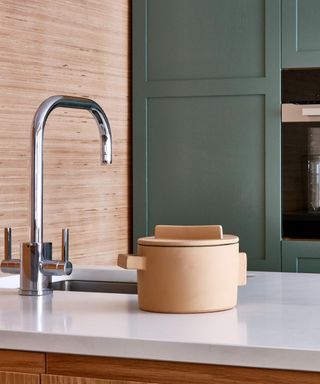 A modern kitchen with sage green cabinetry, a chrome tap, and a white stone worktop. A tan ceramic pot sits on the counter, with textured wood panelling behin