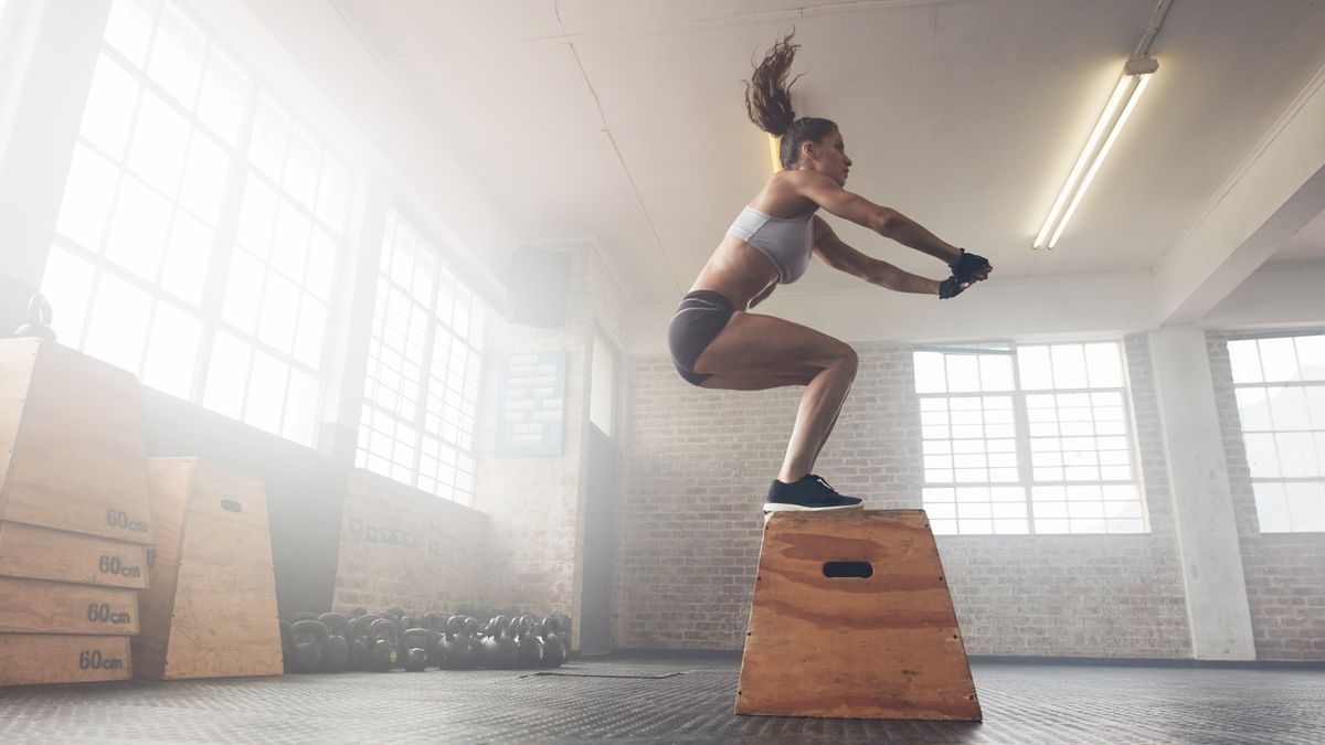 Woman performing a box jump in a warehouse gym landing with both feet on a box during bodyweight workout