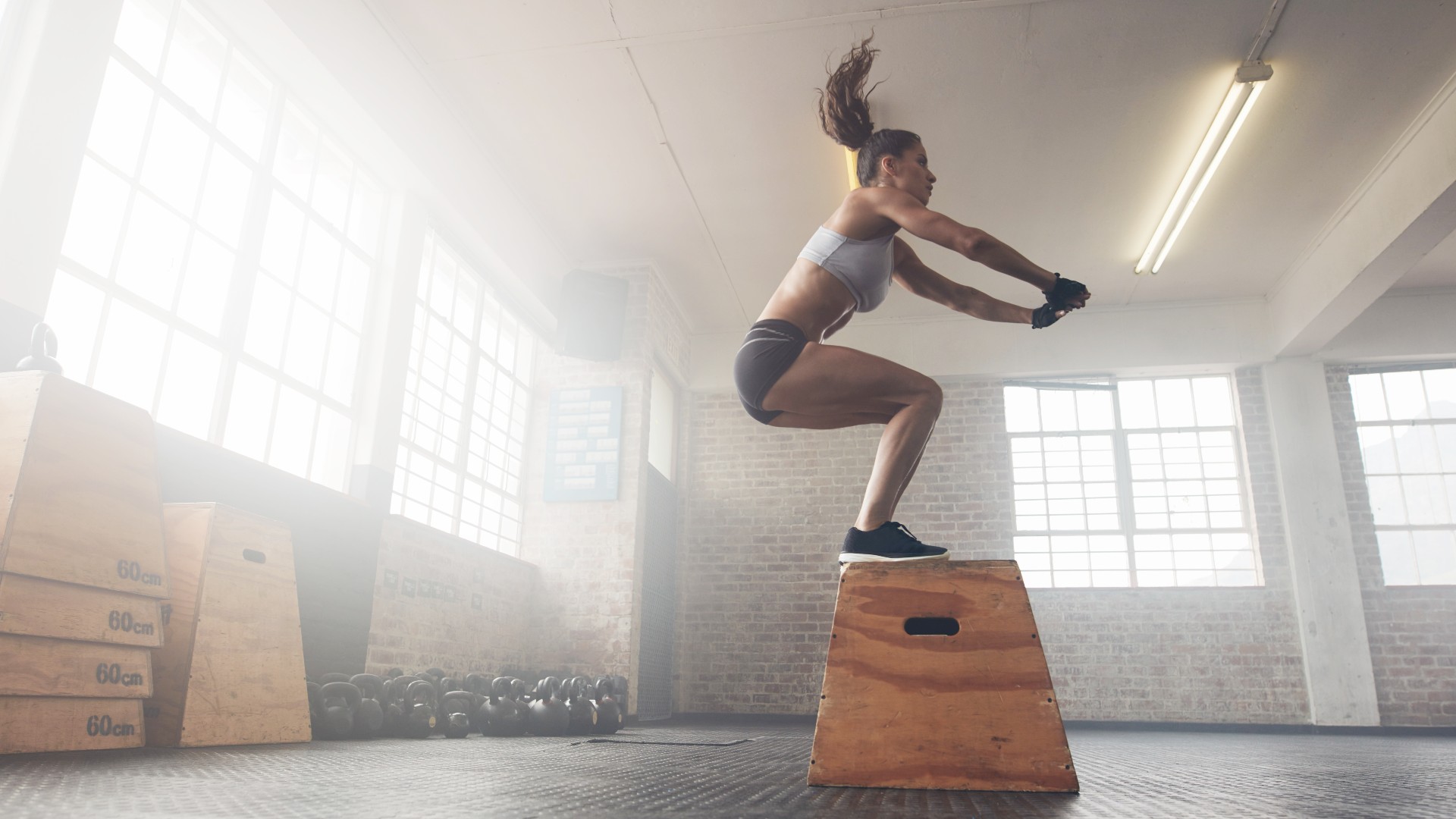 Frau führt in einem Fitnessstudio in einer Lagerhalle einen Boxsprung aus und landet beim Körpergewichtstraining mit beiden Füßen auf einer Box