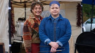 Rochenda Sandall in a brown coat as Fi and Siobhán McSweeney in a blue hat and coat as Della stand in front of a tent in Amandaland.