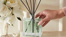 A female holding one of our best reed diffusers (Nest NY Wild Mint & Eucalyptus) with a bouquet of white peonies in background