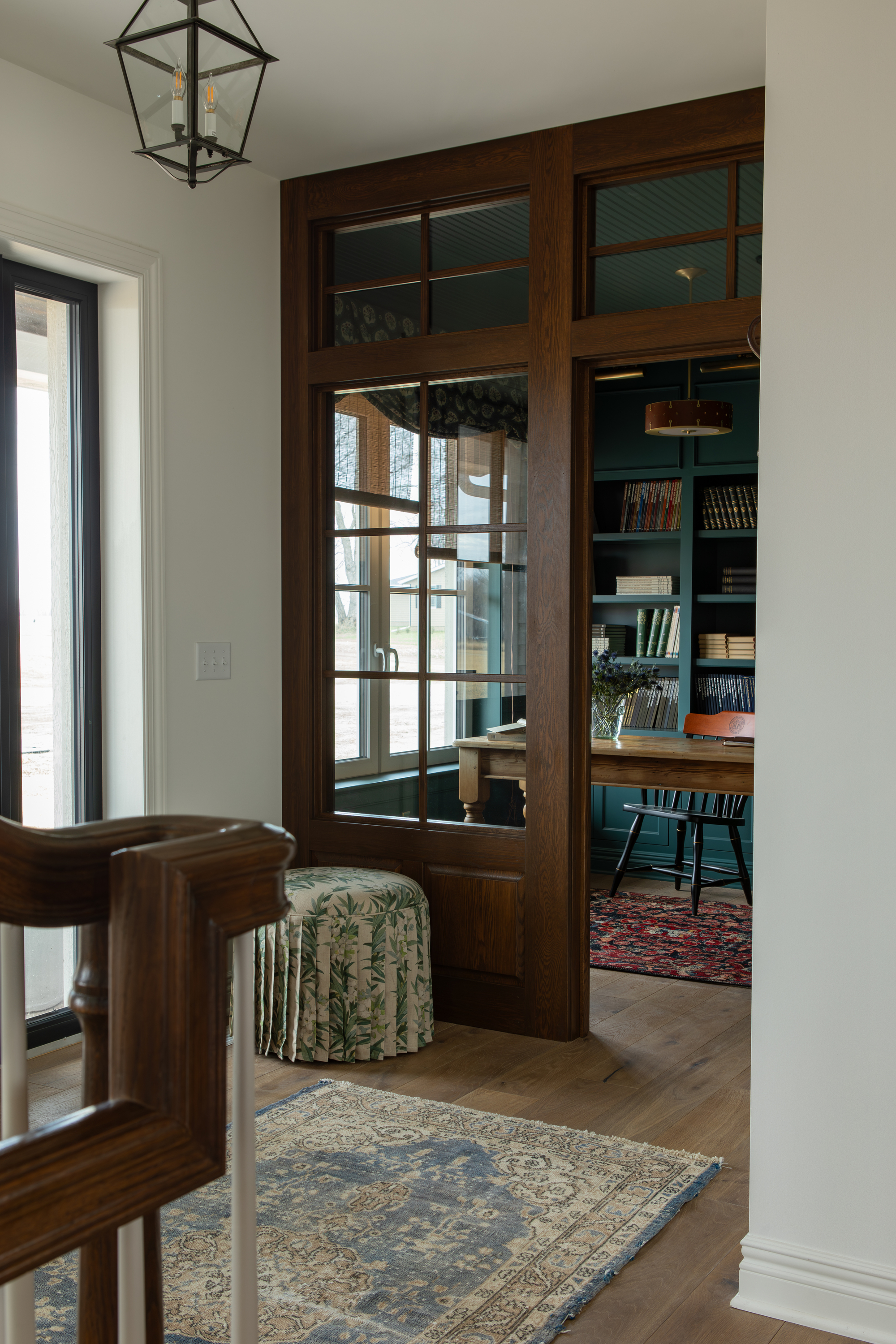 Image of a study with floor-to-ceiling bookshelves painted in a dark blue/green as seen from a white entryway. The entryway is white, and the door to the study is made of dark wood-framed windows.