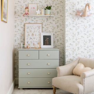 Corner of a nursery with white floral wallpaper, a striped accent chair and a light sage chest of drawers